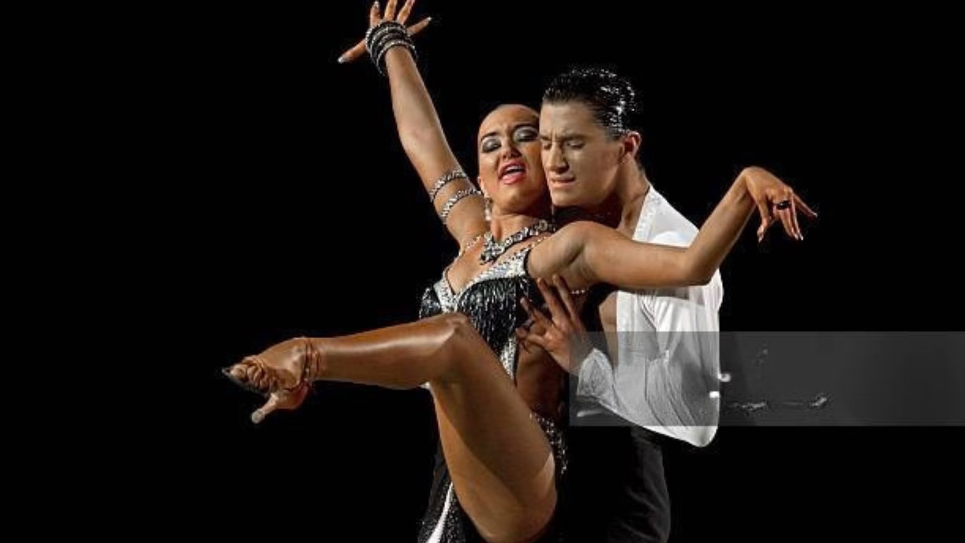 A Latin dance couple in a dramatic performance pose against a black stage.